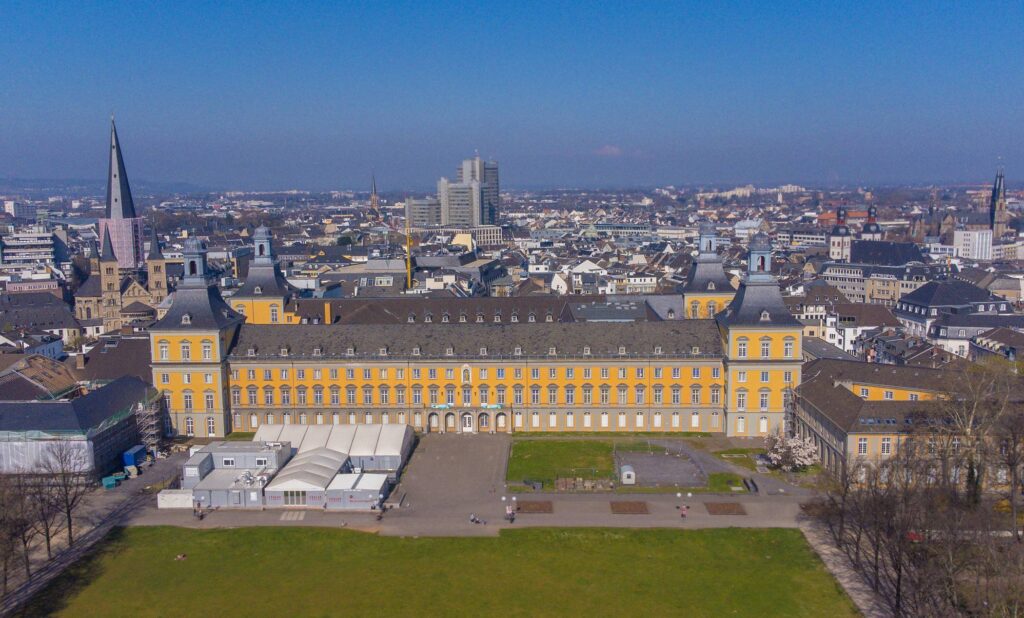 Panoramic aerial view of the University of Bonn’s historic yellow building, set against the backdrop of the city and a clear blue sky.